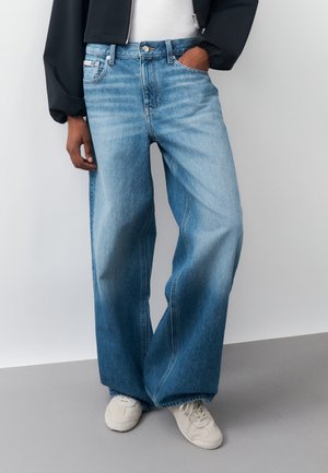 Person iført blå jeans med brede ben og gradientfarvning samt hvide sneakers, med hånden i lommen, foran en ensfarvet lys baggrund.