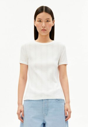 Jeune femme aux cheveux raides foncés porte un haut en maille blanc à manches courtes et un jean bleu clair taille haute, debout devant un fond blanc.