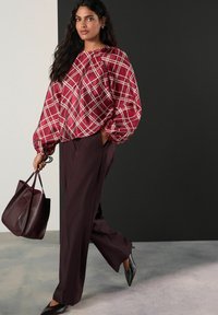 Kvinde går i bordeauxfarvede brede bukser, rødternet puffærmet bluse, sorte spidse sko og bærer en stor mørk bordeauxfarvet taske.