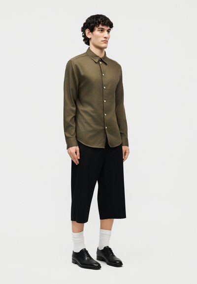 Νέος άνδρας με σγουρά μαλλιά που φοράει πουκάμισο χακί με κουμπιά, μαύρο φαρδύ παντελόνι μέχρι τη γάμπα, λευκές κάλτσες και μαύρα επίσημα παπούτσια, όρθιος ακίνητος.