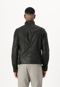 Black leather jacket with a high collar, textured finish, prominent seams, and side buckles; worn over gray trousers.