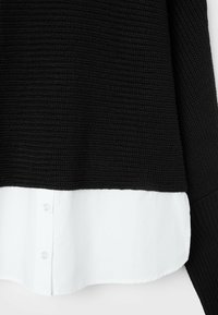 Pull noir tricoté superposé à une chemise blanche avec des boutons visibles en bas de l'ourlet.
