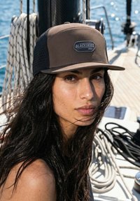 Mujer con cabello largo y oscuro que lleva una gorra marrón y negra con la etiqueta "BLACKSKIES PRO" en la cubierta de un barco con cuerdas enrolladas y el océano al fondo.