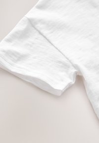 Fehér pamut póló, puha textúrával. A képen a hüvely szélét és a varrás részleteit láthatjuk közelről.