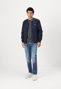 Veste de bombardier marine avec fermeture éclair, poignets côtelés et un petit logo sur la poitrine ; associée à une chemise grise et un jean bleu clair.