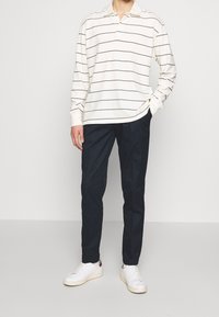 Koszula polo z długim rękawem w kolorze off-white z czarnymi poziomymi pasekami, zestawiona z ciemnonawigacyjnymi spodniami i białymi trampkami. Luźny krój i styl.