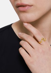 Anello ondulato in oro con finitura liscia e lucida. Mano poggiata sotto il mento, con un colore naturale delle unghie. Sfondo scuro.