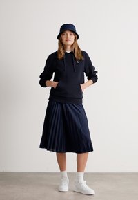 Mujer con un sombrero de cubo negro, sudadera negra con capucha, falda midi azul marino con plisados, calcetines blancos y zapatillas blancas de pie contra un fondo liso.