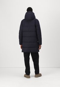 Marineblå polstret parka med hætte, der har horisontale quiltede sektioner og en lige pasform. Kombinationen med mørke bukser og lysebrune sko.