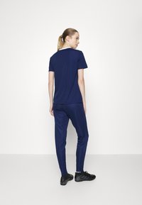 Námornícky modrý športový outfit pozostávajúci z trička s krátkym rukávom a zúžených nohavíc, vyrobený z hladkej látky. Čierne športové topánky dopĺňajú vzhľad.