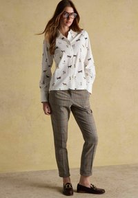 Žena oblečená v bielej košeli s gombíkmi a potlačou psov, károvaných nohaviciach po členky, s veľkými okuliarmi a tmavými mokasínami so zlatými reťazovými detailmi.