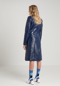 Cappotto trapuntato blu navy con finitura lucida, maniche lunghe e design di lunghezza media. Indossato con calzini blu a fantasia e tacchi bianchi.