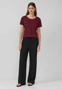 T-shirt bordeaux à manches courtes avec un col rond associé à un pantalon large noir à texture lâche. La tenue est complétée par des sandales noires.
