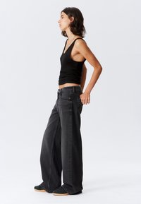 Giovane donna in top nero, jeans neri a gamba larga sbiaditi e sneakers scure, in profilo su uno sfondo chiaro e semplice.