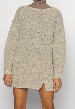 Longue robe-pull beige en maille avec une fente latérale, portée sur un col roulé marron, montrée du cou jusqu'à la mi-cuisse.