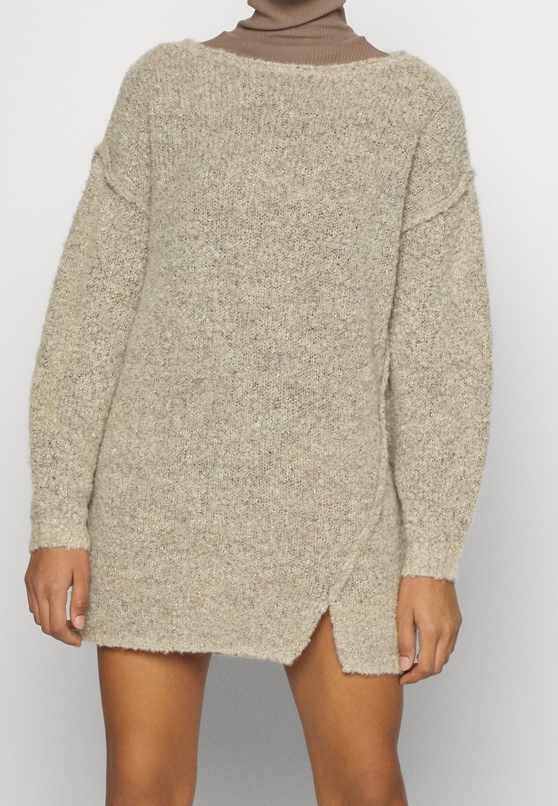 Longue robe-pull beige en maille avec une fente latérale, portée sur un col roulé marron, montrée du cou jusqu'à la mi-cuisse.