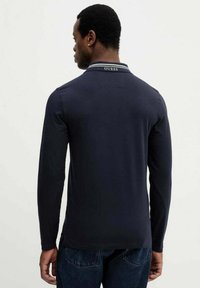 Granatowa koszulka polo z długim rękawem, białym kołnierzykiem w paski i logo "GUESS" na plecach. Wykonana z gładkiej bawełnianej tkaniny.