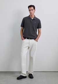 Camisa polo gris con cuello en contraste, pantalones blancos y zapatos negros. La tela es suave. La camisa presenta un emblema en el pecho.