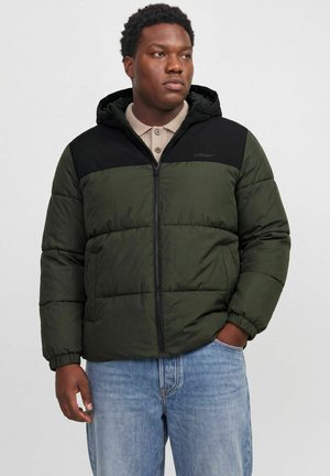 Homme portant une doudoune verte et noire avec capuche, une chemise beige boutonnée et un jean bleu, debout avec une main dans la poche.