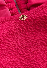 Fuchsia-farbener strukturierter Stoff mit einem gewebten Muster. Verfügt über elastische Raffungen und einen goldfarbenen runden Verschluss mit quadratischem mittlerem Detail.