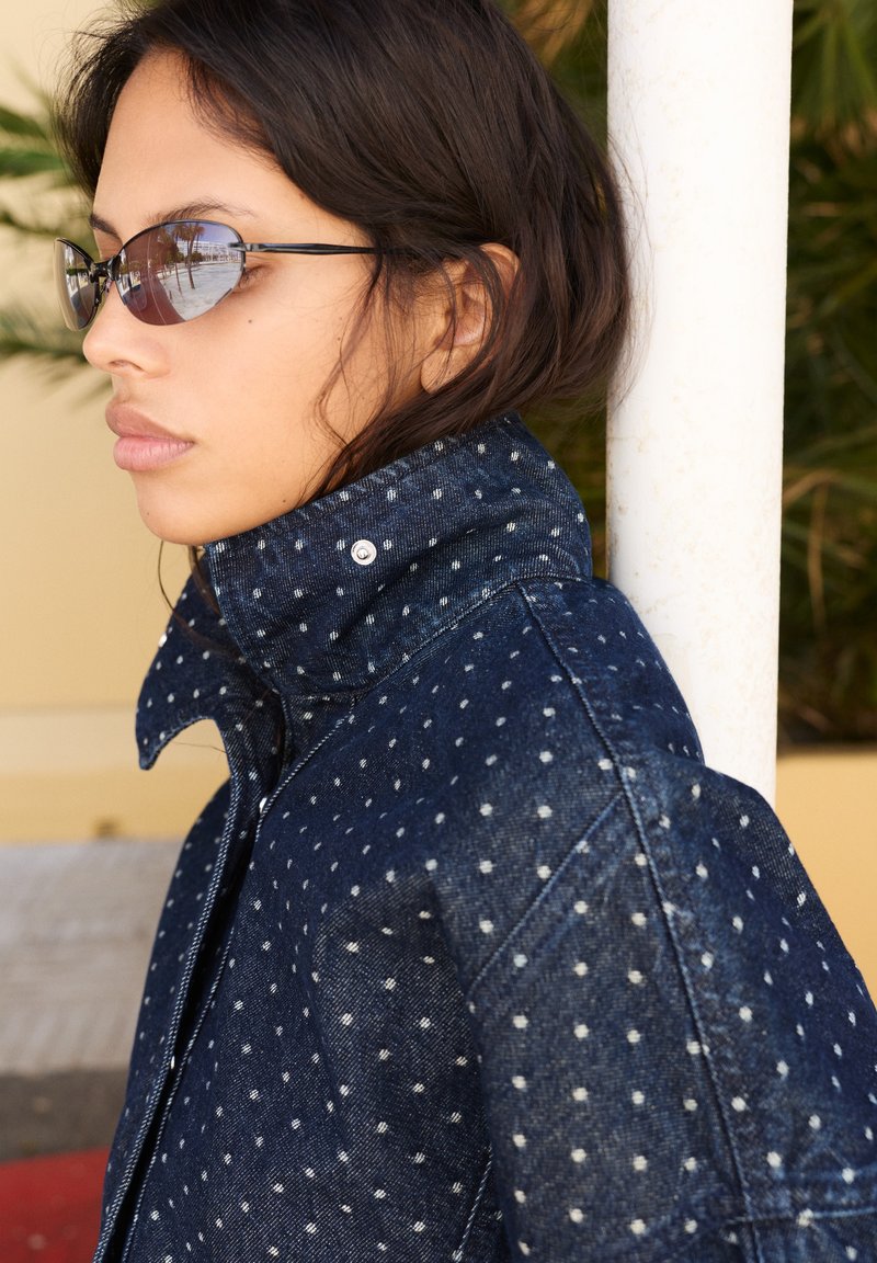 Jeune personne aux cheveux courts et foncés portant de petites lunettes de soleil ovales et une veste en jean bleu foncé à pois blancs, tournée vers la gauche.