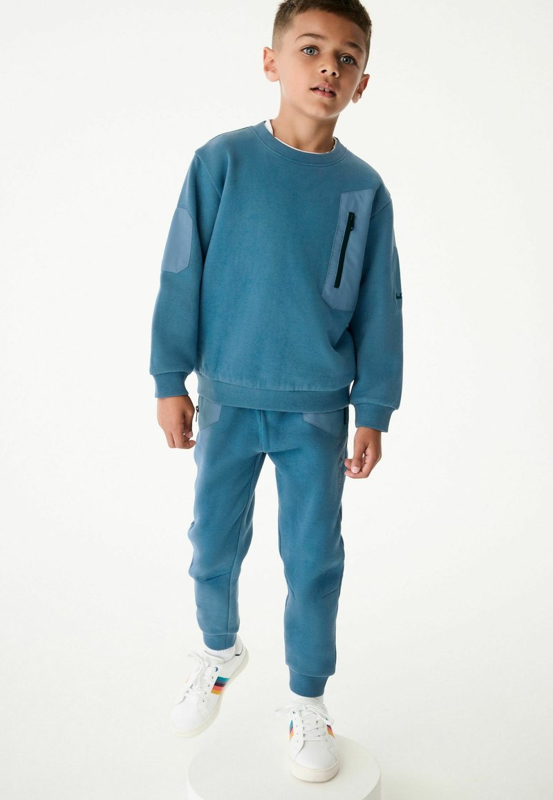 Felpa blu e pantaloni abbinati in morbido tessuto; presenta una tasca con zip, polsini a costine e dettagli a pannello sulle maniche. Sneakers bianche con accenti di colore.