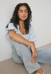 Hellblaues Kurzarmoberteil mit Puffärmeln, kombiniert mit hochgeschnittenen hellen Jeans. Das Model posiert sitzend mit lockigem Haar und natürlichem Make-up.