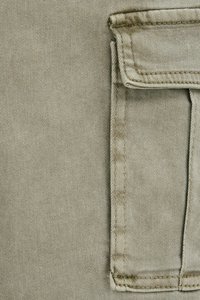 Detailní záběr na béžovou tkaninu s obdélníkovou klopovou kapsou, která má viditelné stehy a přehnutý okraj na pravé straně.