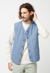 Hellblauer gesteppter Weste mit einem Knopfverschluss vorne, die vertikale Nähte und runde Kanten aufweist, getragen über einem beigen Pullover.