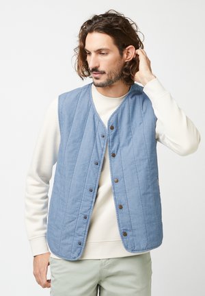 Light blue quilted vest with a button-front closure, featuring vertical stitching and round edges, worn over a beige sweater.