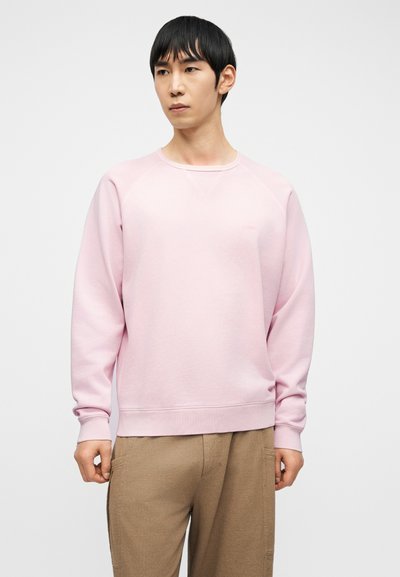 Jeune homme aux cheveux noirs et raides portant un sweat-shirt rose clair et un pantalon beige, debout devant un fond clair uni.