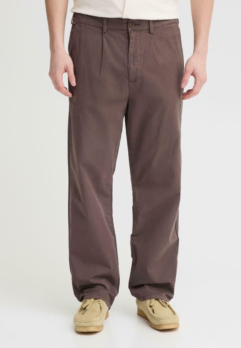 Homme portant un pantalon marron foncé à coupe ample et des mocassins beiges, debout devant un fond clair uni.