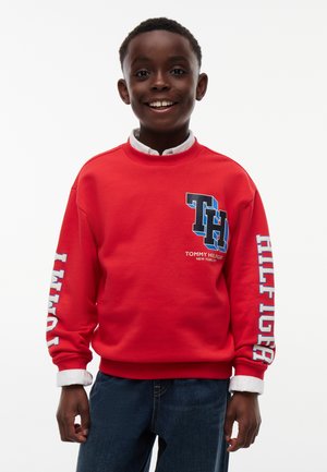 Jonge jongen glimlacht en draagt een rode Tommy Hilfiger trui met logo op de borst en tekst op de mouwen, gecombineerd met een blauwe spijkerbroek.