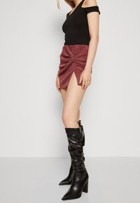 Mujer con un top negro de hombros al descubierto, una falda mini arrugada rosa brillante con abertura frontal y botas altas negras con tacón y estilo holgado, de pie de lado.