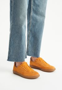 Pomarańczowe zamszowe sneakersy z płaskimi sznurowadłami, gumową podeszwą i subtelnymi detalami szycia, noszone z jasnoniebieskimi dżinsami z postrzępionymi końcami.