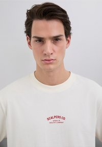 Sudadera color crema con cuello redondo, con un logotipo bordado en rojo que dice "SCALPERS CO." y el texto "SPORTY & HEALTHY COMPANY" en el pecho.