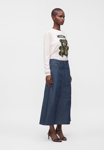 Femme debout portant un sweat-shirt blanc Moschino avec un motif ours en peluche, une longue jupe en jean bleue et des sandales noires à talons hauts.