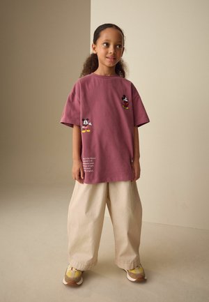 Ragazza giovane con capelli ricci che indossa una maglietta larga color bordeaux di Topolino, pantaloni beige larghi e scarpe da ginnastica gialle, in piedi all'interno.