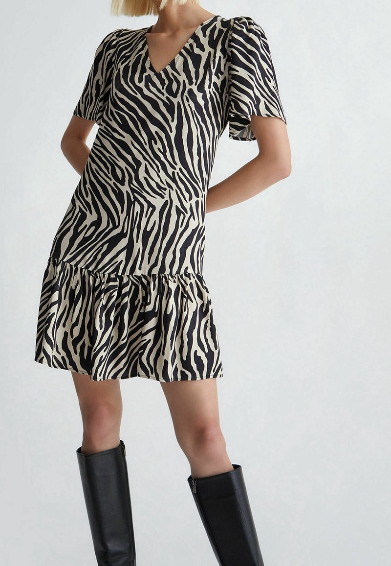 Robe à imprimé zèbre noir et beige avec des manches courtes bouffantes, un décolleté en V et un ourlet à volants, associée à des bottes noires ajustées jusqu'aux genoux.