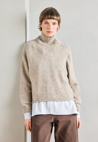 Béžový pletený svetr s vysokým límcem, texturovaná tkanina a pružné manžety, vrstvený přes světle modrou košili s límcem a hnědé kalhoty.