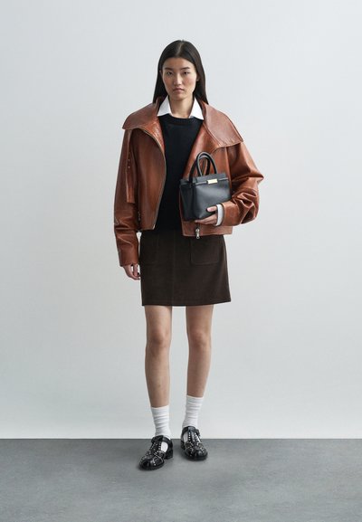 Brun læder oversized jakke, sort sweater, brun nederdel, sort håndtaske, hvide sokker, sorte sko med nitter, stående mod en lys baggrund.