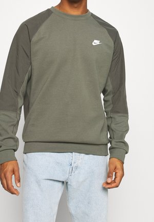 Mężczyzna w oliwkowej bluzie Nike z ciemnozielonymi rękawami i jasnoniebieskich dżinsach, stojący na białym tle.