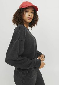 Sudadera negra de corte relajado y hombros caídos. Tejido texturizado y puños acanalados. Combinada con una gorra roja.