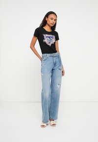 Kvinne i sort grafisk t-skjorte, lyseblå høytlivs ripped jeans med belte, og beige åpen-tå hæler som står mot hvit bakgrunn.