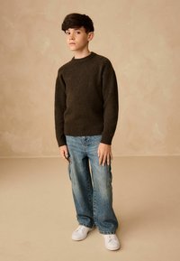 Brązowy sweter o teksturze, niebieskie luźne dżinsy, białe sneakersy. Stoi na beżowym tle. Naturalne światło podkreśla detale.
