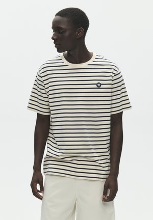 Mężczyzna w kremowo-granatowej koszulce w paski z krótkim rękawem i jasnych spodniach, patrzący na lewo na jednolitym tle.