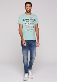 Mátově zelené tričko s potiskem v černé a oranžové barvě. Nošeno s vybledlými modrými džíny a bílými teniskami. Model stojí a usmívá se.