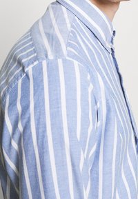 Mężczyzna w jasnoniebieskiej i białej koszuli z pionowymi paskami, zapinanej na guziki, z kołnierzykiem, przedstawiony od ramienia do dolnej części twarzy.