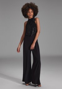 Czarny top halter z cekinami i drapowanym dekoltem, zestawiony z szerokimi czarnymi spodniami. Modelka stoi na neutralnym tle.