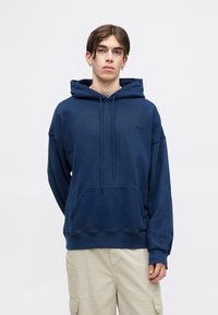 Granatowy hoodie wykonany z miękkiego materiału, z kieszenią typu kangur, kapturem ze sznurkiem oraz ściągaczami przy mankietach i hemie. Prosty detal z logo na klatce piersiowej.